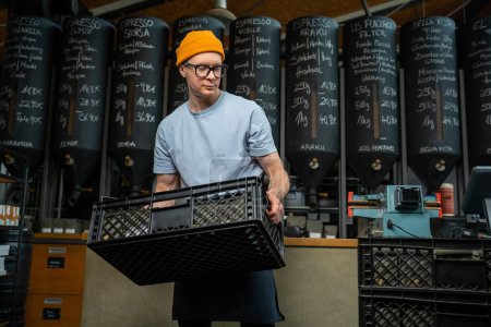 Photo for Caucasian man wearing hat apron and glasses coffee shop worker gets new batch of coffee packages in boxes on roasting factory. - Royalty Free Image