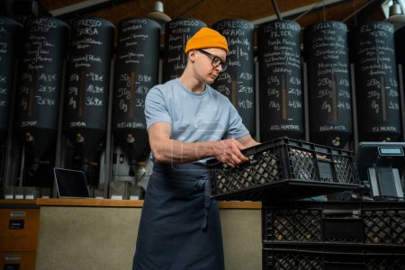 Téléchargez les photos : Homme caucasien portant tablier chapeau et lunettes cafetière travailleur obtient nouveau lot de paquets de café dans des boîtes sur l'usine de torréfaction. - en image libre de droit