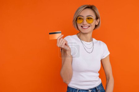 Foto de Encantadora mujer feliz sonriente en camiseta blanca que muestra la banca con tarjeta de crédito aislada sobre fondo naranja. - Imagen libre de derechos