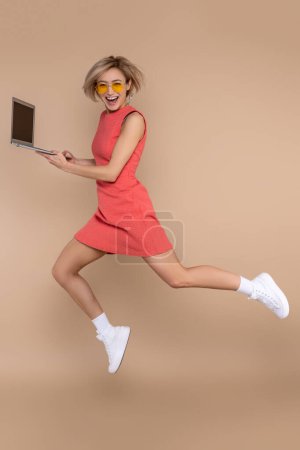 Foto de Mujer saltando en vestido y gafas de sol usando portátil trabajando en línea aislado sobre fondo beige. - Imagen libre de derechos
