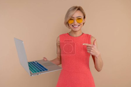 Photo for Delighted woman in dress and sunglasses pointing at laptop display isolated over beige background. - Royalty Free Image