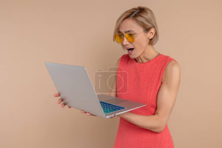 Foto de Mujer sorprendida en vestido y gafas de sol mirando a la pantalla del ordenador portátil con la boca abierta trabajando en línea aislado sobre fondo beige. - Imagen libre de derechos