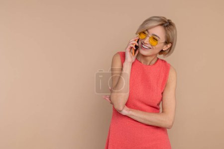 Foto de Feliz mujer sonriente en vestido y gafas de sol hablando de teléfono móvil aislado sobre fondo beige. - Imagen libre de derechos