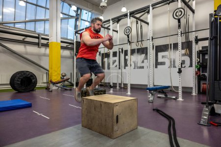 Photo for Caucasian bearded man having workout, jumping up on wooden box in crossfit gym. - Royalty Free Image