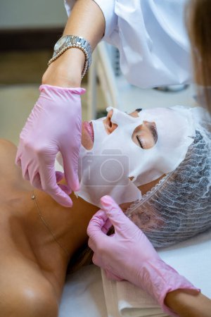Foto de Esteticista irreconocible aplicación de mascarilla de hoja en la cara de la joven cliente. - Imagen libre de derechos