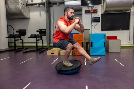 Photo for Fit athletic man performing exercise on gymnastic hemisphere bosu ball in gym, standing on one leg. - Royalty Free Image