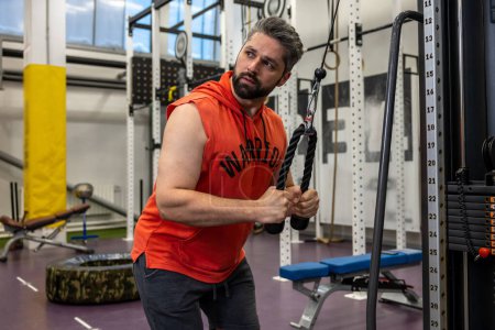 Foto de Hombre adulto barbudo haciendo ejercicio en el gimnasio, haciendo flexiones de cable, bíceps y tríceps entrenamiento de brazo. - Imagen libre de derechos