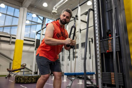 Foto de Hombre adulto barbudo haciendo ejercicio en el gimnasio, haciendo flexiones de cable, bíceps y tríceps entrenamiento de brazo. - Imagen libre de derechos