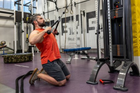 Foto de Hombre deportivo fuerte en tríceps de entrenamiento de ropa deportiva y bíceps en el gimnasio, haciendo empujes de cuerda de cable de pie sobre las rodillas. - Imagen libre de derechos