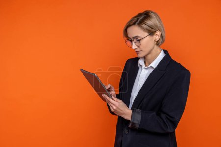 Foto de Mujer seria que usa traje negro de estilo oficial que trabaja en línea en la tableta aislada sobre fondo naranja. - Imagen libre de derechos