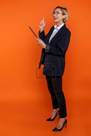 Téléchargez les photos : Délicieuse femme en costume noir de style officiel levant le doigt ayant l'idée de travailler en ligne sur tablette isolée sur fond orange. - en image libre de droit