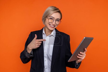 Téléchargez les photos : Femme heureuse portant un costume de style officiel noir pointant vers la tablette isolée sur fond orange. - en image libre de droit