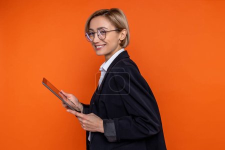 Téléchargez les photos : Portrait de profil de femme heureuse portant costume de style officiel noir travaillant en ligne sur tablette isolée sur fond orange. - en image libre de droit