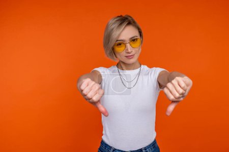 Photo for Displeased woman in white t shirt showing dislike gesture thumbs down isolated over orange background. - Royalty Free Image