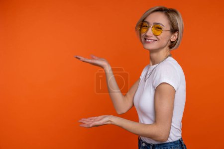 Foto de Perfil retrato de mujer en camiseta blanca mostrando área para publicidad con las manos de gran tamaño aislado sobre fondo naranja. - Imagen libre de derechos