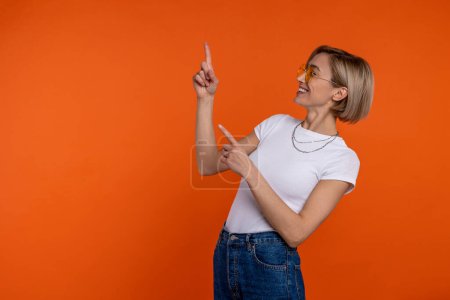 Foto de Mujer feliz en camiseta blanca y jeans apuntando hacia arriba en el espacio de copia, mostrando el área de publicidad aislada sobre fondo naranja. - Imagen libre de derechos