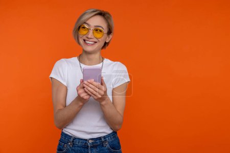 Téléchargez les photos : Femme positive portant un T-shirt blanc utilisant un téléphone portable vérifiant les réseaux sociaux isolés sur fond orange. - en image libre de droit
