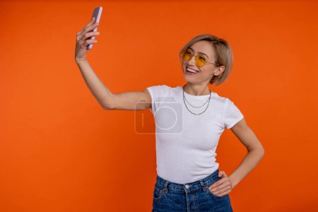 Foto de Blogger mujer satisfecha en camiseta blanca y jeans haciendo selfie en smartphone aislado sobre fondo naranja. - Imagen libre de derechos