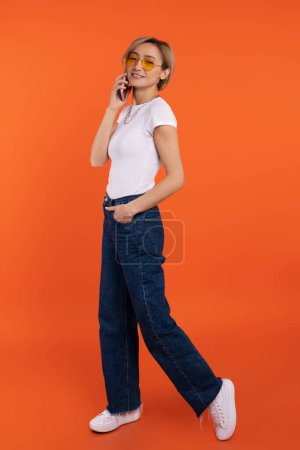 Foto de Hermosa mujer en camiseta blanca y pantalones vaqueros hablando en el teléfono móvil aislado sobre fondo naranja. - Imagen libre de derechos
