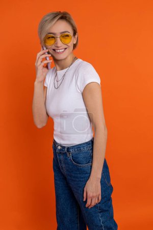 Foto de Mujer satisfecha en camiseta blanca y jeans hablando en teléfono móvil aislado sobre fondo naranja. - Imagen libre de derechos