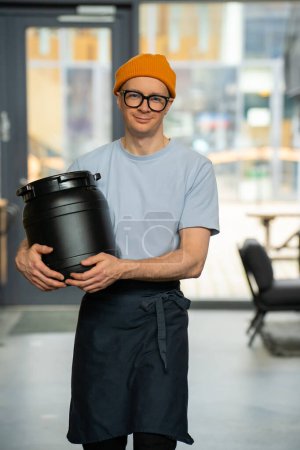 Téléchargez les photos : Jeune barista hipster portant chapeau, lunettes et tablier portant tonneau se préparant au travail dans un café. - en image libre de droit