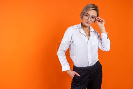 Foto de Mujer seria vistiendo camisa blanca de estilo oficial sosteniendo gafas mirando hacia otro lado espacio libre aislado sobre fondo naranja. - Imagen libre de derechos