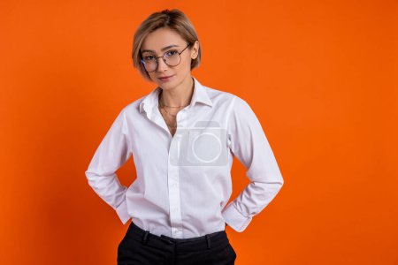 Foto de Mujer ambiciosa con camisa blanca de estilo oficial de pie con las manos en las caderas aisladas sobre fondo naranja. - Imagen libre de derechos