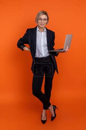 Foto de Hermosa mujer con traje negro de estilo oficial que sostiene el ordenador PC aislado sobre fondo naranja. - Imagen libre de derechos
