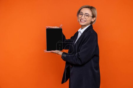 Foto de Mujer alegre sonriente con traje negro de estilo oficial que muestra la pantalla vacía de la tableta aislada sobre fondo naranja. - Imagen libre de derechos