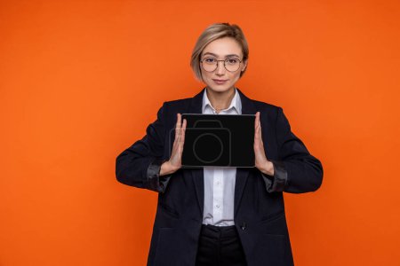 Téléchargez les photos : Femme confiante portant costume de style officiel noir montrant écran vierge de comprimé isolé sur fond orange. - en image libre de droit