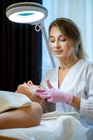 Photo for Cosmetologist apply cream on womans face, caucasian client woman in beauty salon get spa procedures, cleansing face. - Royalty Free Image