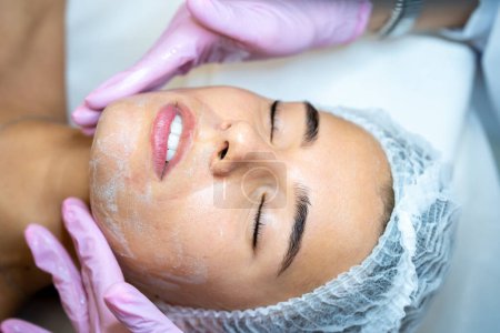 Téléchargez les photos : Cosmétologue féminine méconnaissable appliquer un masque blanc sur le visage des femmes, procédures de spa, nettoyage du visage. - en image libre de droit
