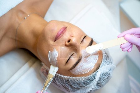Téléchargez les photos : Cosmétologue méconnaissable appliquant le masque à la femme visage dans le salon de beauté spa. - en image libre de droit