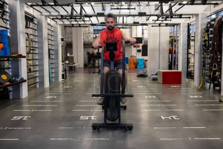 Téléchargez les photos : Homme caucasienportif en vêtements de sport faisant du vélo sur des vélos d'exercice à la salle de gym, entraînement cardio. - en image libre de droit