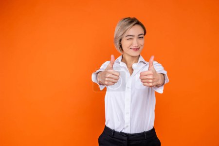 Foto de Mujer positiva con camisa blanca de estilo oficial que muestra como gesto y guiño a la cámara aislada sobre fondo naranja. - Imagen libre de derechos