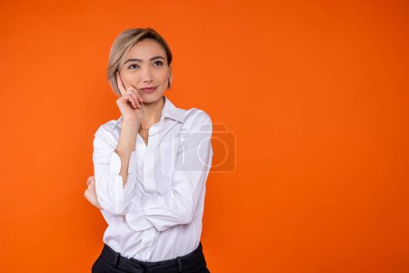 Téléchargez les photos : Femme pensive portant une chemise de style officiel blanc pensant tenant menton espace libre isolé sur fond orange. - en image libre de droit