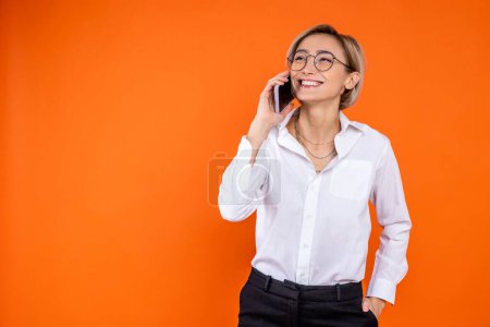 Foto de Mujer sonriente con camisa blanca de estilo oficial hablando en un teléfono inteligente aislado sobre fondo naranja. - Imagen libre de derechos