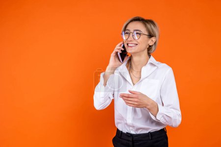 Foto de Mujer satisfecha vistiendo camisa blanca de estilo oficial hablando en teléfono inteligente aislado sobre fondo naranja. - Imagen libre de derechos