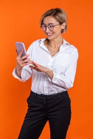 Téléchargez les photos : Joyeux belle femme portant chemise de style officiel blanc en utilisant un téléphone mobile isolé sur fond orange. - en image libre de droit
