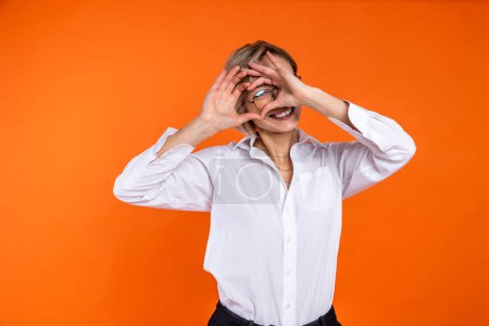 Téléchargez les photos : Femme romantique portant une chemise de style officiel blanc regardant à travers la forme du cœur avec les mains isolées sur fond orange. - en image libre de droit