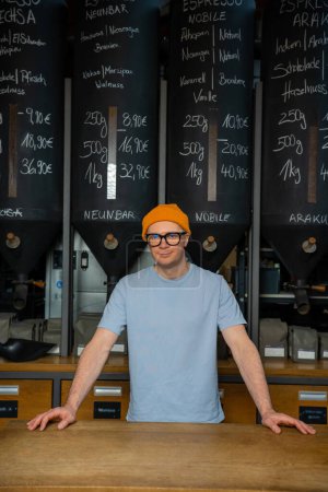 Foto de Guapo barman hipster con gorro sombrero y vasos en el interior de la cafetería moderna con gran variedad de granos de café. - Imagen libre de derechos