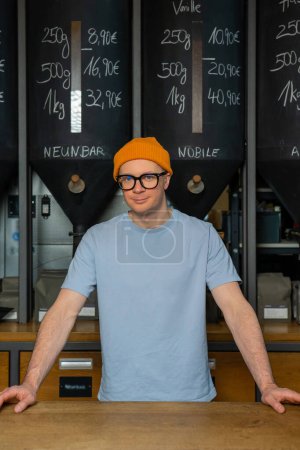 Photo for Friendly hipster bartender wearing beanie hat and glasses in the interior of modern coffee shop with big variety of coffee beans. - Royalty Free Image