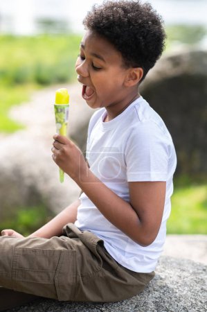 Téléchargez les photos : Mignon petit garçon heureux mangeant de la crème glacée à l'extérieur pendant la chaude journée d'été, assis sur le rocher, savourant un dessert sucré pendant les vacances d'été. - en image libre de droit