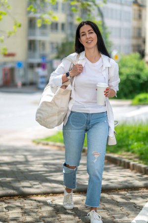 Photo for Beautiful smiling young woman with white bag standing on city street, drinking coffee in paper, waking in town. - Royalty Free Image