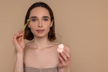 Téléchargez les photos : Femme brune utilisant un rouleau facial pour les procédures de soins de la peau tenant l'éponge cosmétique debout isolé sur fond beige. - en image libre de droit