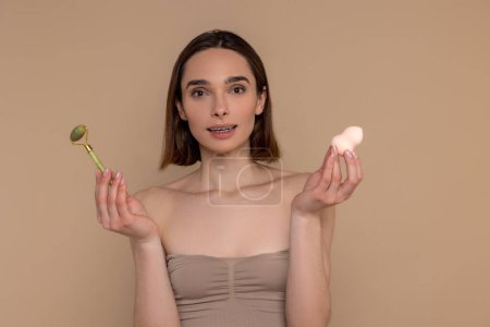 Photo for Brunette woman using facial roller for skin care procedures holding cosmetic sponge standing isolated over beige background. - Royalty Free Image