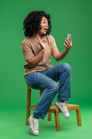 Photo for Connected. Excited young woman sitting on the chair with a phone in hands - Royalty Free Image
