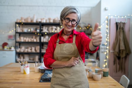 Photo for Smiling senior woman potter master posing in her workshop among earthenware products showing like gesture, thumb up. - Royalty Free Image