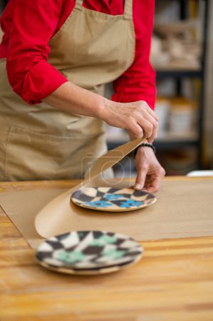 Téléchargez les photos : Mains de maître de poterie de femme enveloppant l'article de faïence commandé par le client dans le papier dans le lieu de travail - en image libre de droit