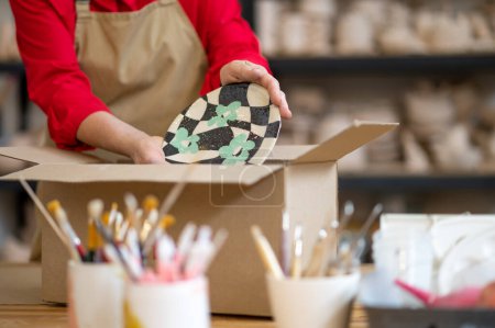 Téléchargez les photos : Femme travailleuse méconnaissable de la commande d'emballage de magasin de poterie pour le client tout en se tenant à table contre les étagères. - en image libre de droit